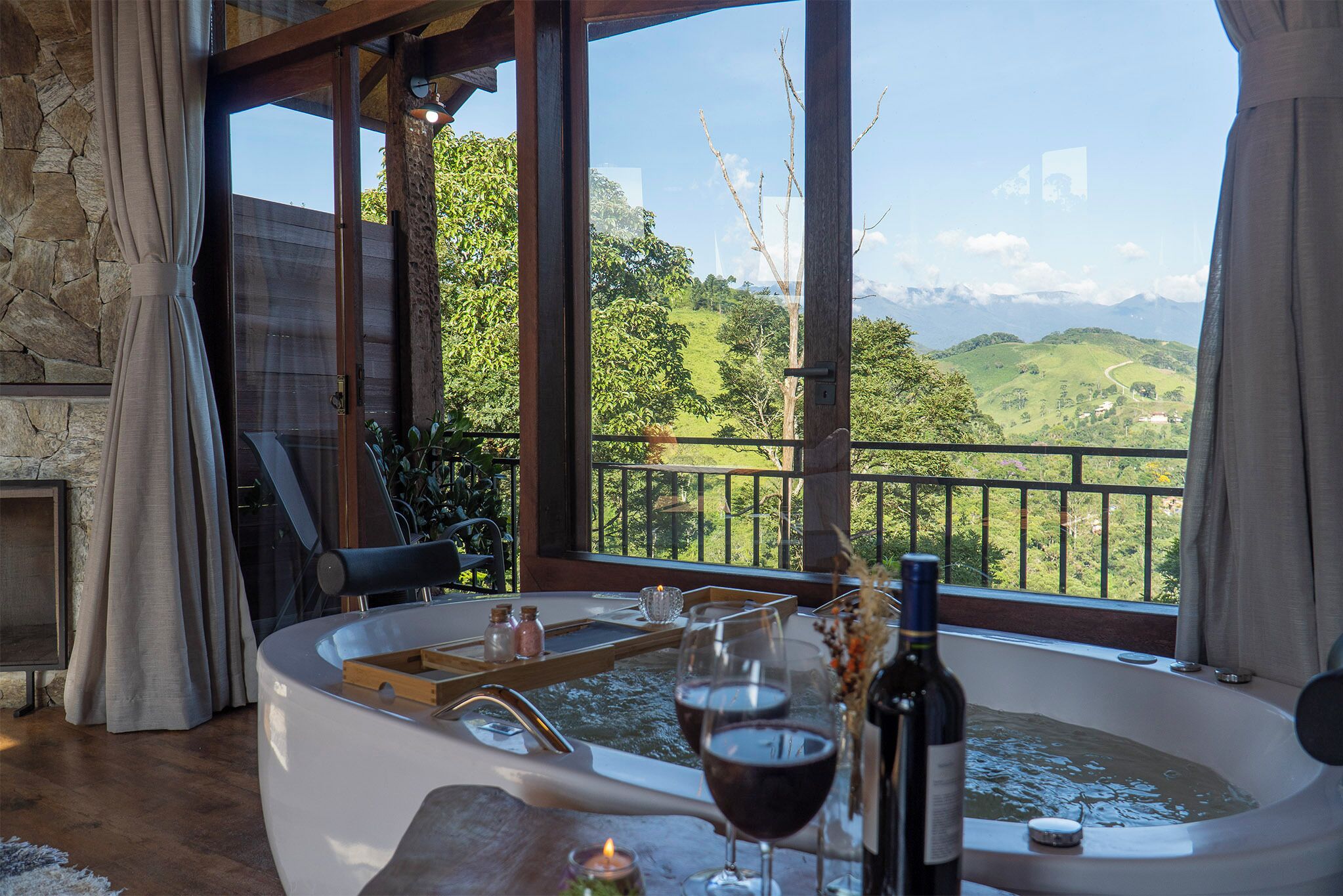A room with a large bathtub, a wooden table, and a view of green hills.