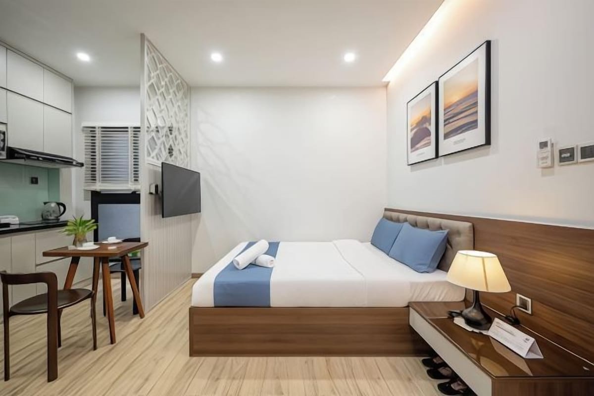 A modern hotel room with a bed, bedside table, lamp, and a small desk with a chair.