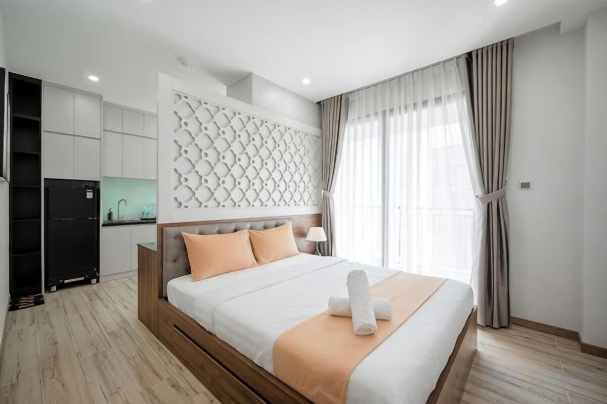 A modern hotel room with a large bed, a built-in headboard, a kitchenette, and a view of the city.