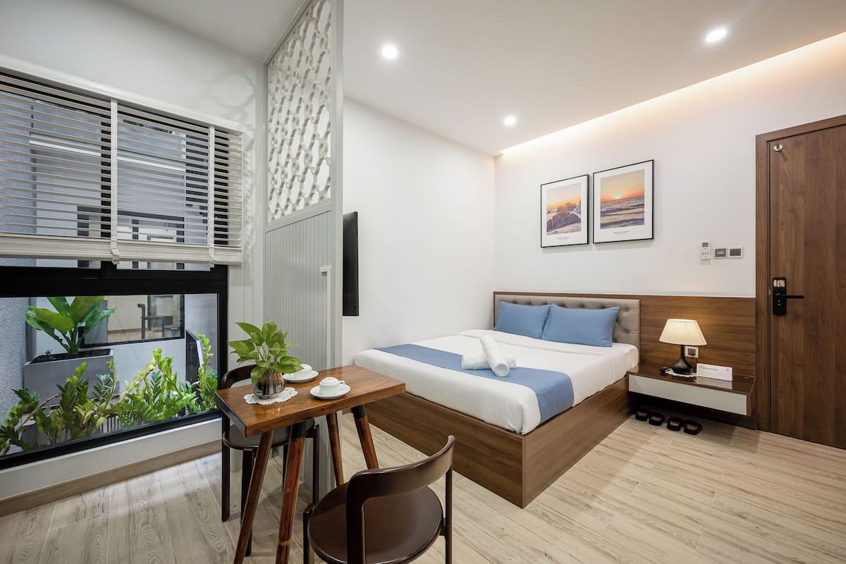 A modern hotel room with a large bed, a wooden nightstand, a desk with a chair, and a window with blinds.