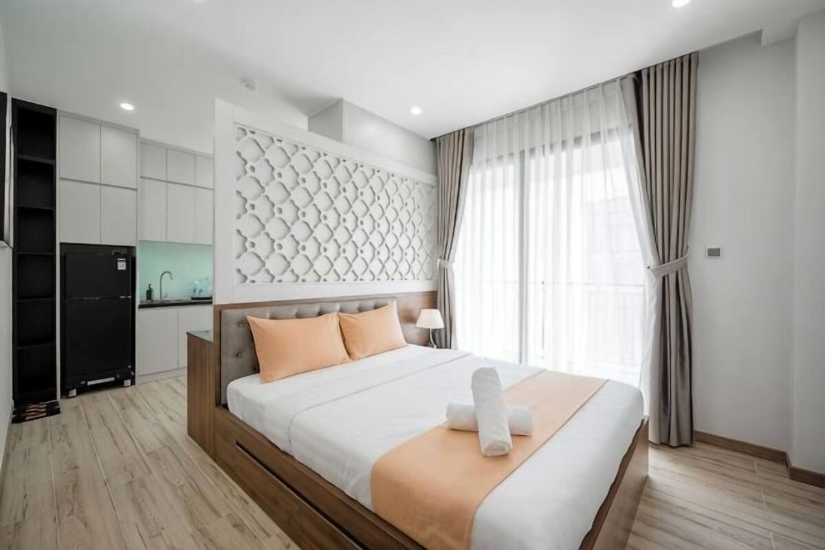 A modern hotel room with a large bed, a built-in wardrobe, a kitchenette, and a large window with curtains.