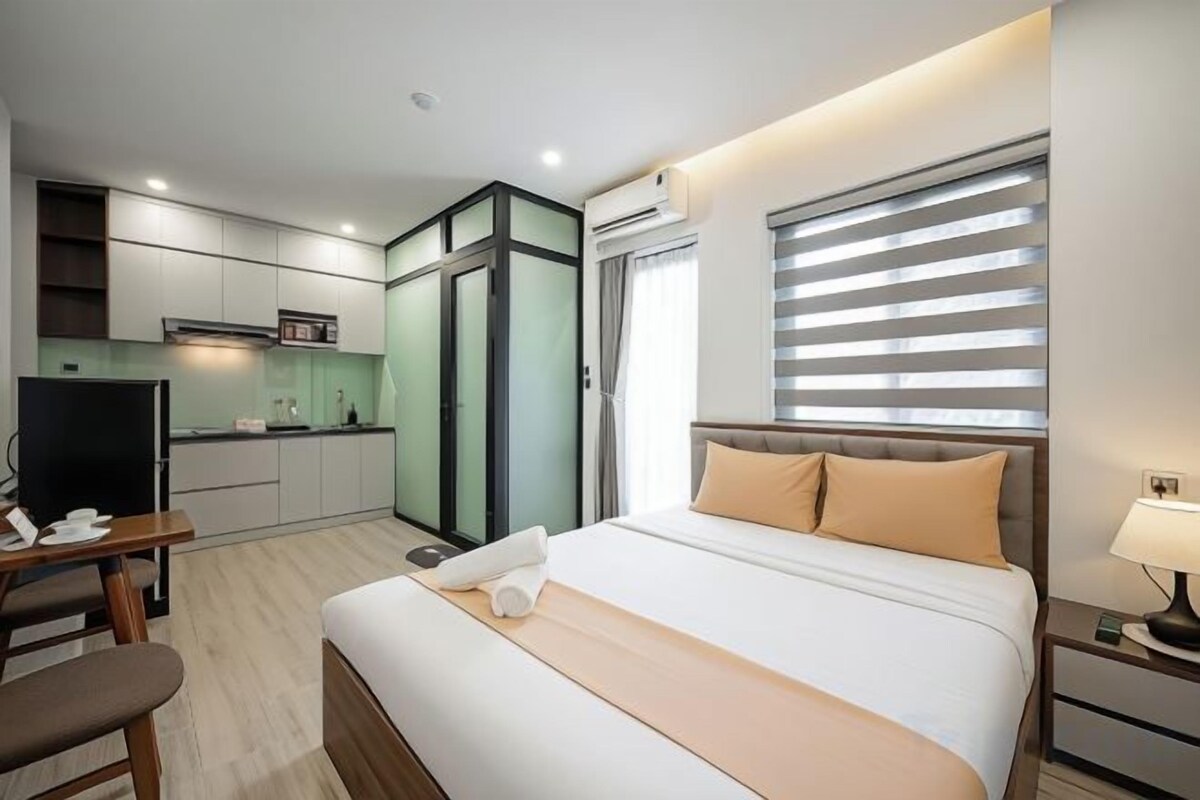 A hotel room with a bed, bedside table, a desk, a chair, a TV, a kitchenette, and a window with blinds.