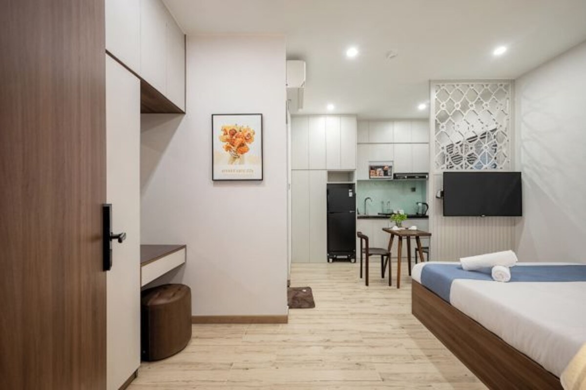 A modern hotel room with a bed, a desk, a chair, a TV, and a kitchenette.