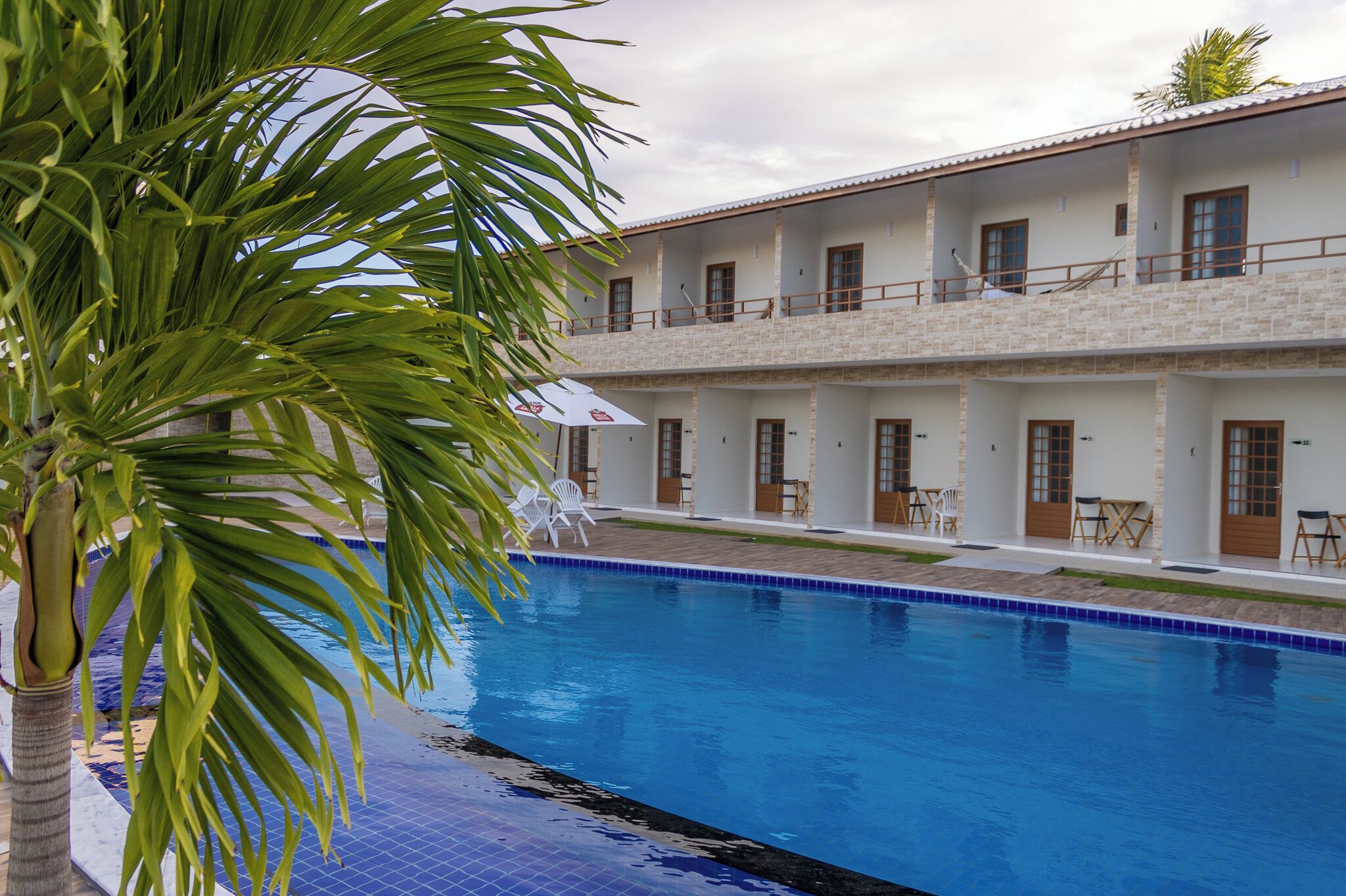 A poolside view of a hotel with a two-story building, outdoor seating, and a palm tree.