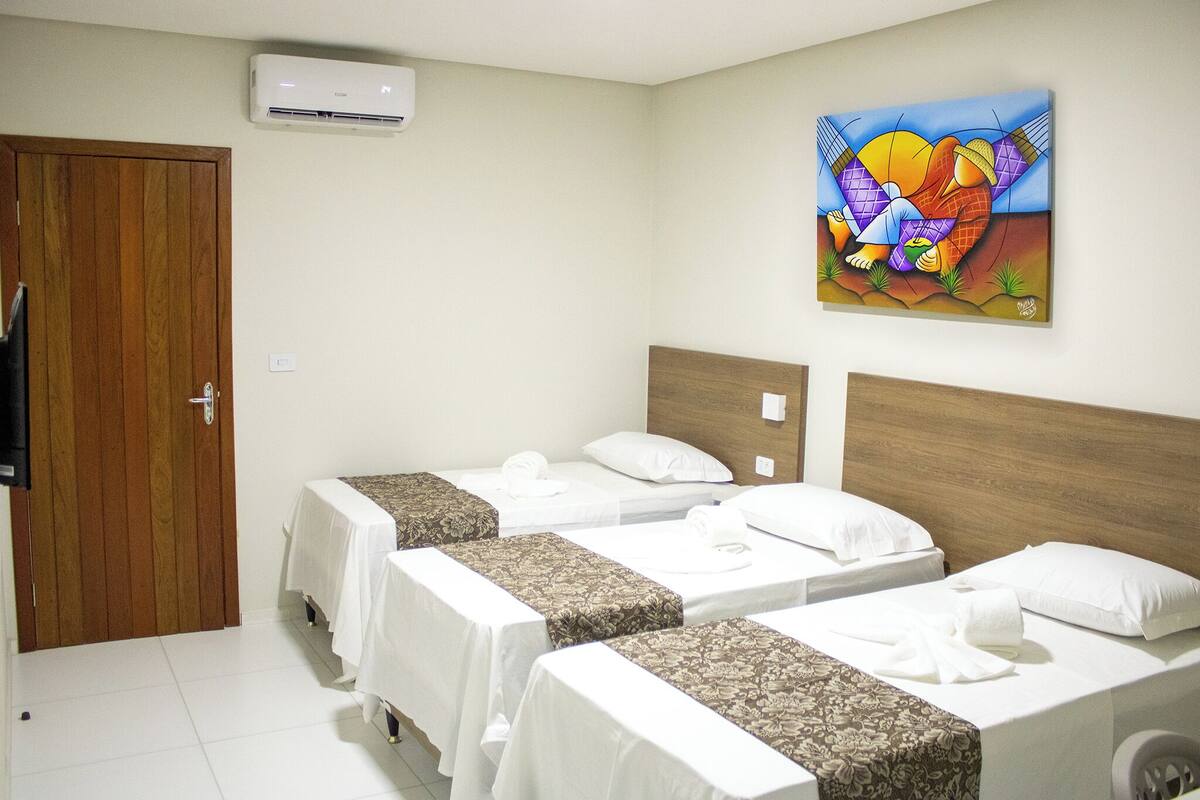A hotel room with three single beds, a wooden door, an air conditioning unit, and a colorful painting on the wall.