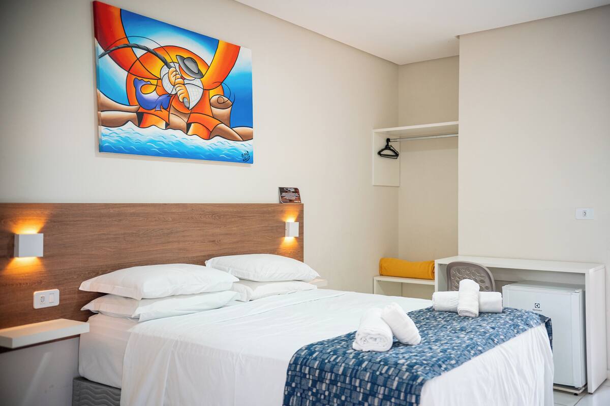 A hotel room with a bed, a desk, a chair, a small refrigerator, and a painting on the wall.