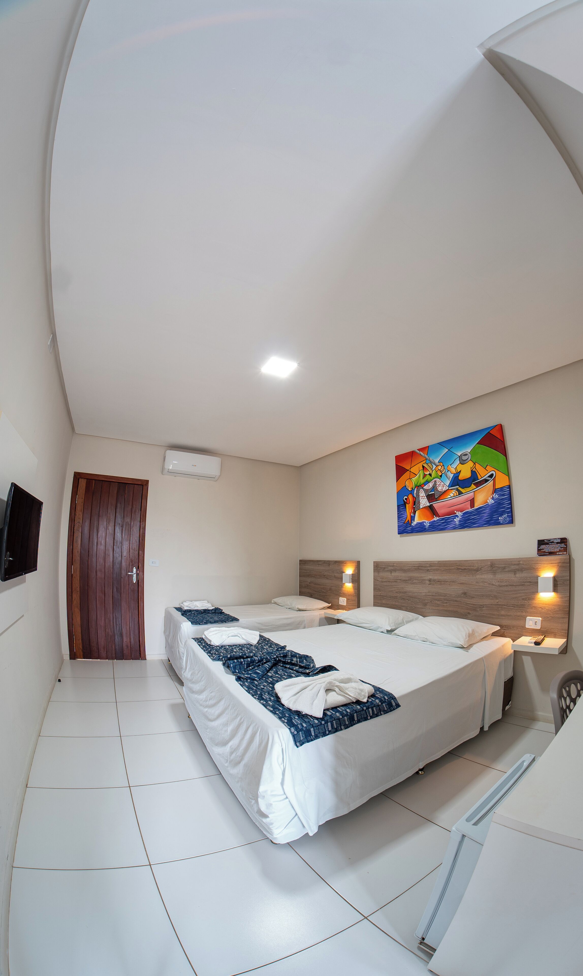 Habitación de hotel con dos camas, una puerta de madera, un equipo de aire acondicionado, un cuadro y una mesita con lámpara.