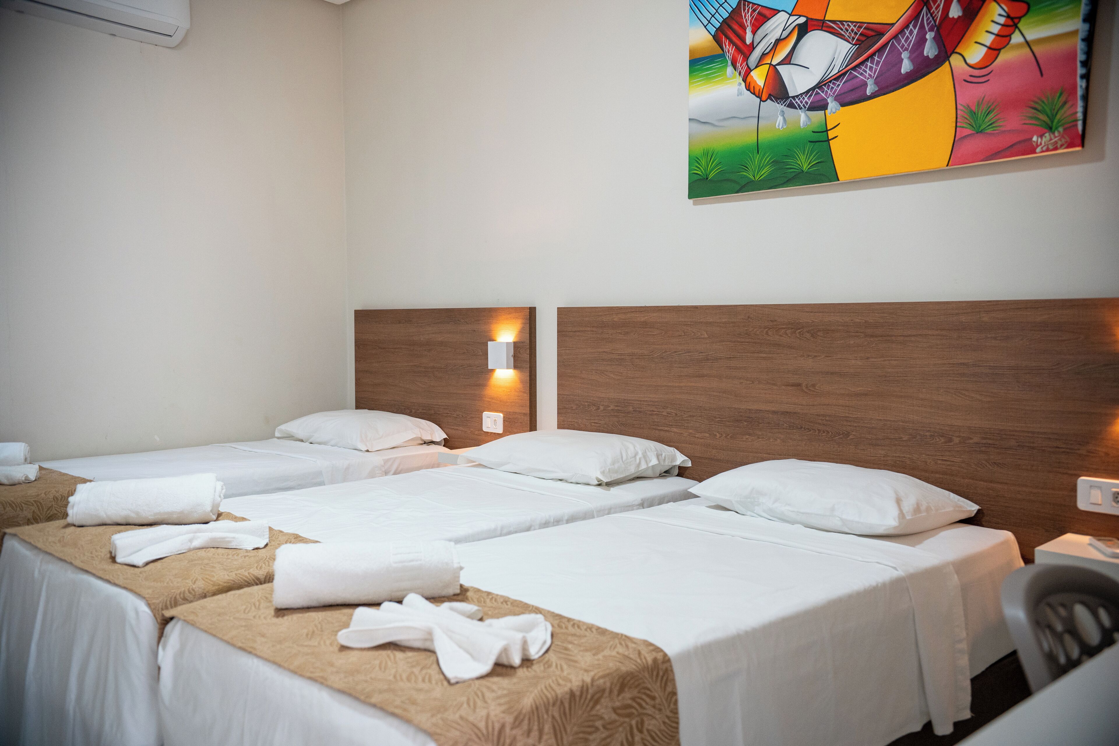 Quarto de hotel com três camas de solteiro, cada uma com lençóis brancos e cabeceira marrom. Há uma pintura abstrata colorida na parede acima das camas.