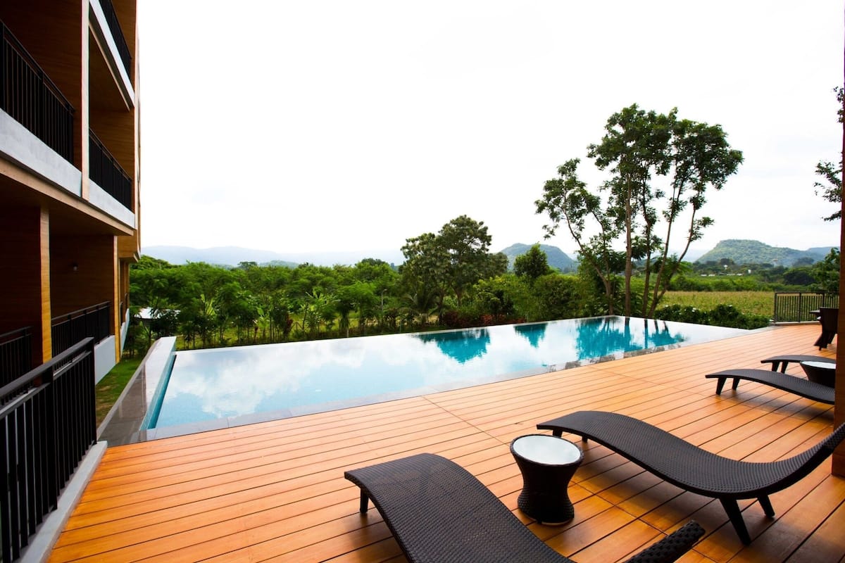 An infinity pool with wooden deck and lounge chairs, surrounded by lush greenery and hills.