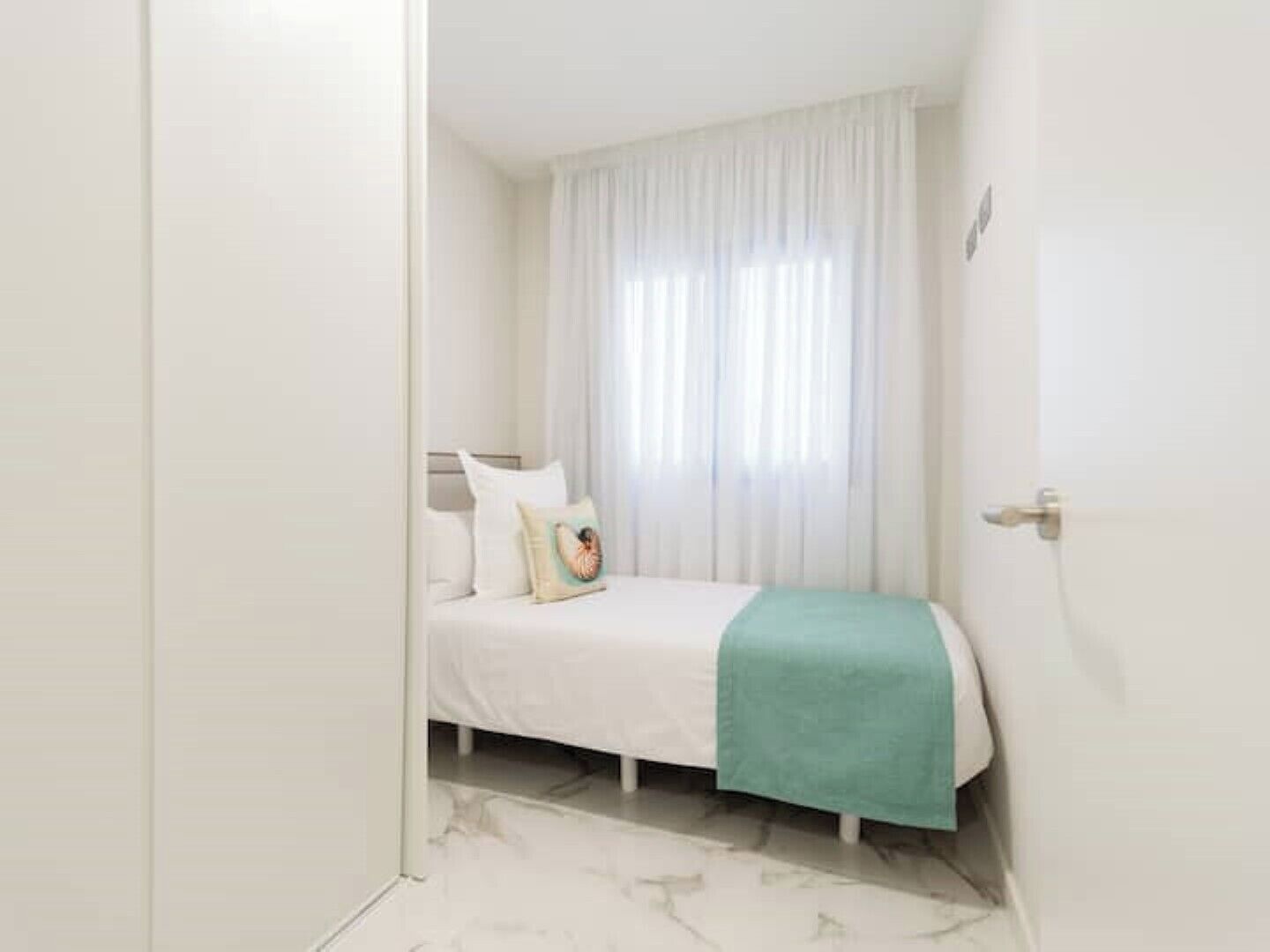 Une petite chambre propre, avec un lit, des rideaux blancs et une porte.