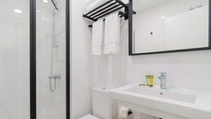 A modern bathroom with a glass shower enclosure, a white toilet, a sink with a mirror above it, and a wooden shelf with two white hats.