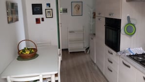 Kapea keittiö, jossa on valkoiset kaapit, liesi ja vihreä, seinälle ripustettu pannunalusta. Keittiössä on valkoinen pöytä tuoleineen ja kori täynnä kukkia.