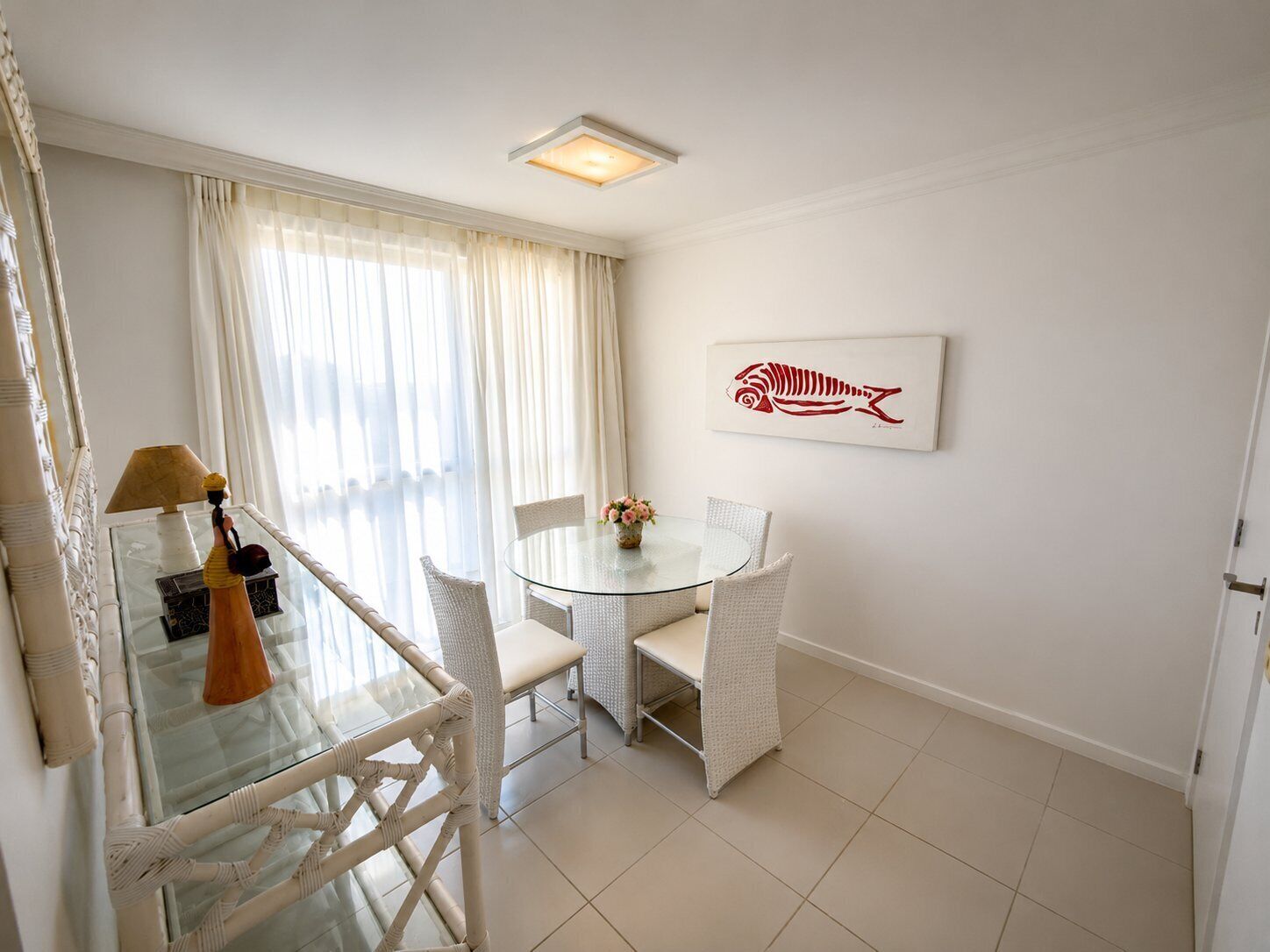 Una habitación con una mesa redonda de vidrio, sillas de mimbre blanco, un jarrón con flores, una lámpara y una obra de arte con motivos marinos en la pared.