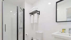 Et moderne badeværelse med brusekabine i glas, toilet, vask med toiletartikler og en træskammel med sutsko.