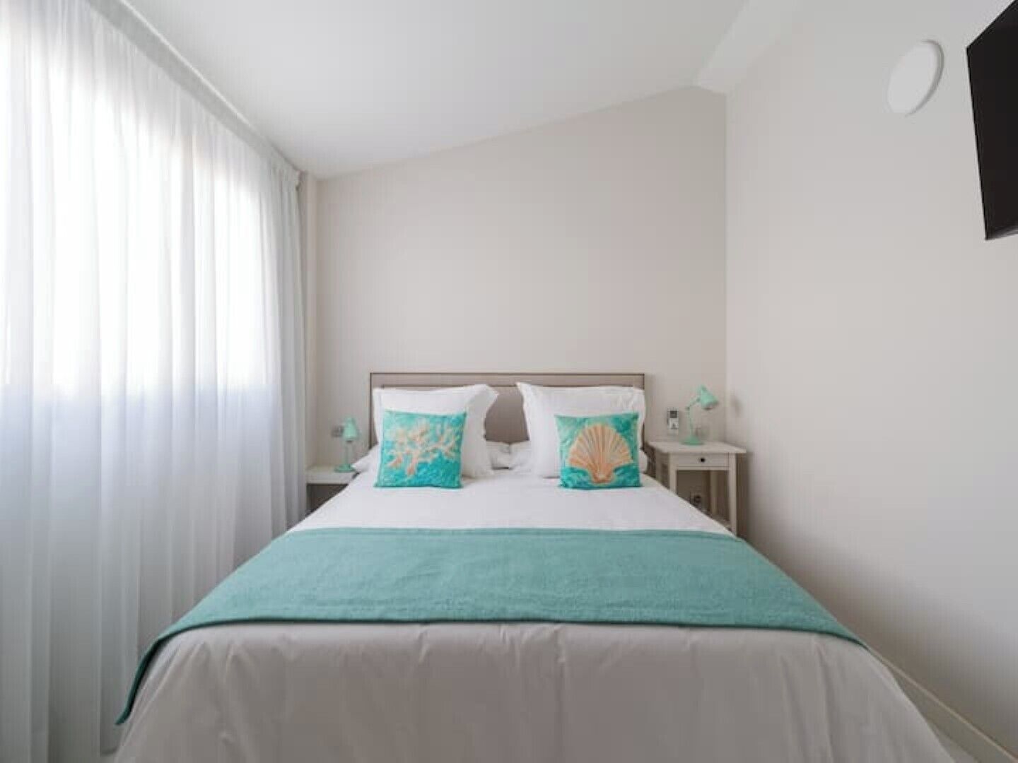 A neatly made bed with a teal bedspread and two decorative pillows, a bedside table with a lamp, and a window with sheer curtains.