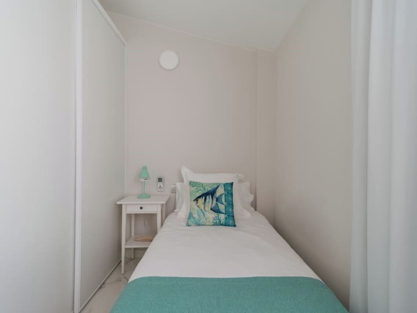Une petite chambre à coucher blanche, avec un lit simple, une table de chevet, une lampe et un luminaire fixé au mur.