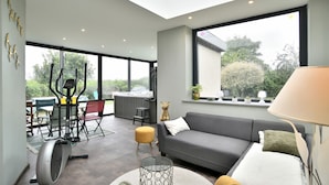 A modern living room with a grey sofa, a standing exercise machine, and a dining area with chairs and a table. - Chez Marie-Céline - Maison avec jacuzzi à 20 min du Val André (Plestan)
