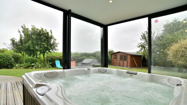 A hot tub in a covered outdoor area with large windows, a wooden deck, and a view of a house and garden. - Chez Marie-Céline - Maison avec jacuzzi à 20 min du Val André (Plestan)