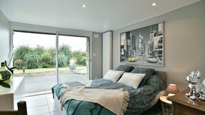 A bedroom with a bed, a table, and a chair. There is a large window and a painting on the wall. - Chez Marie-Céline - Maison avec jacuzzi à 20 min du Val André (Plestan)