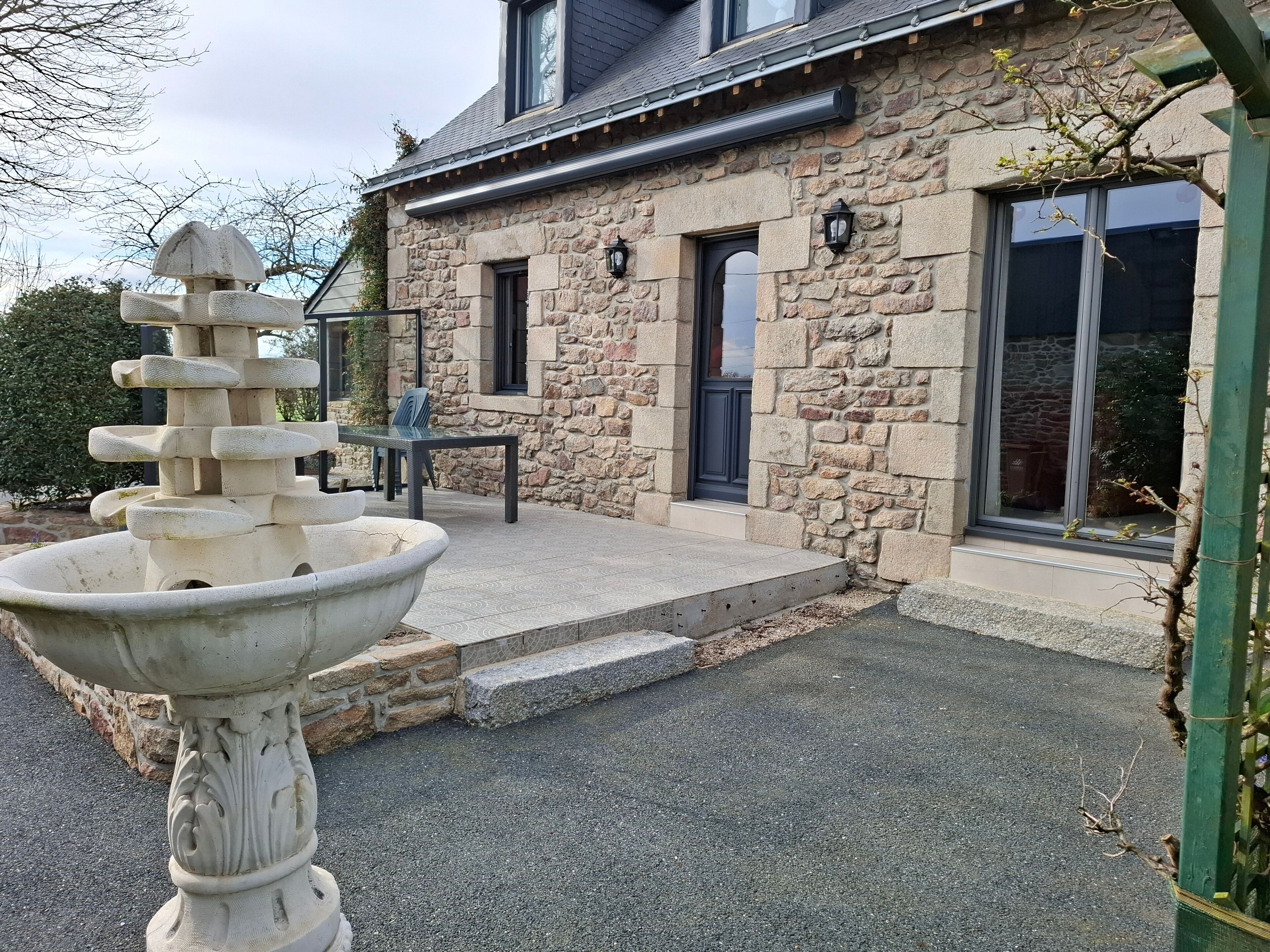 Un bâtiment en pierre avec une fontaine en pierre au premier plan, une table avec des chaises et une porte.