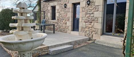 Un bâtiment en pierre avec une fontaine en pierre au premier plan, une table avec des chaises et une porte.