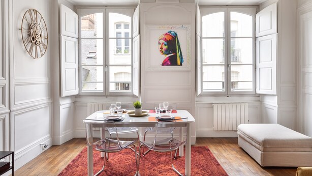 Un comedor con una mesa blanca, sillas transparentes y una alfombra roja. Se observa una ventana con vista a edificios.