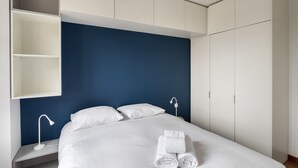 Una habitación de hotel moderna con una cama grande, ropa blanca impecable, una toalla doblada con cuidado, una lámpara de noche y un armario.