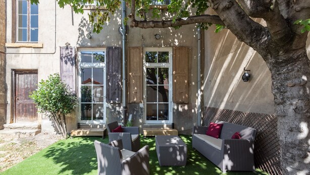 A courtyard with wicker furniture and a tree, surrounded by a building with large windows and shutters. - Le Jardin des Docks - Appt avec jardin (Lyon)