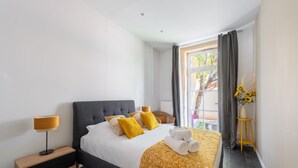 A modern bedroom with a grey upholstered bed, a wooden nightstand, a yellow patterned throw blanket, and a small table with a vase of sunflowers. - Le Jardin des Docks - Appt avec jardin (Lyon)