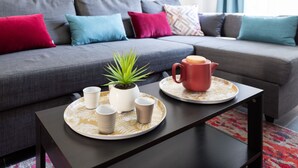 A modern living room with a grey sofa, colorful cushions, a small table with a tray holding cups and a teapot, and a potted plant. - Le Jardin des Docks - Appt avec jardin (Lyon)