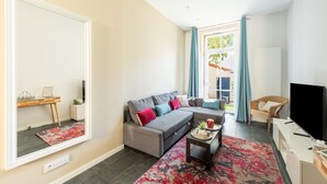 A modern living room with a grey sofa, a coffee table, and a TV. There is a large mirror on one wall and a window with curtains. - Le Jardin des Docks - Appt avec jardin (Lyon)