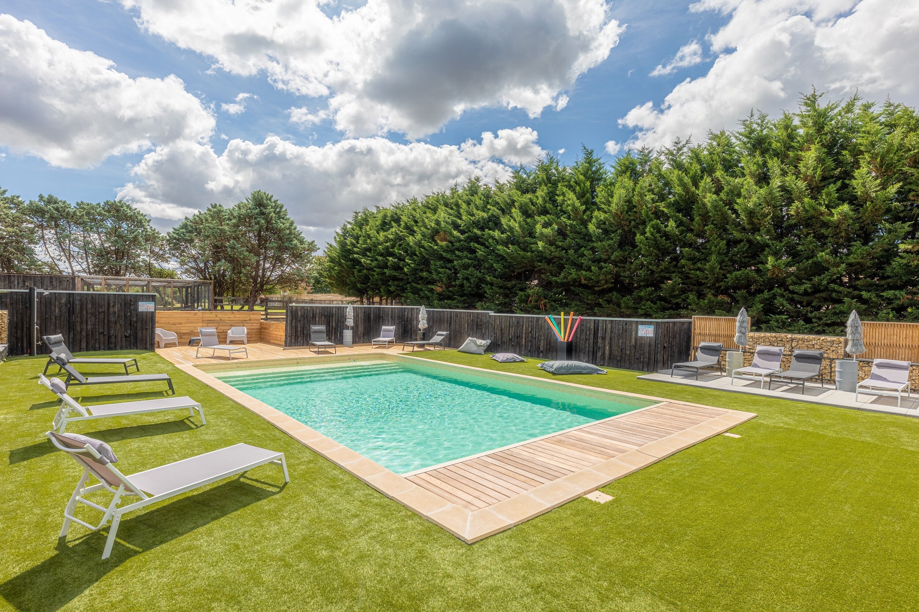 A rectangular swimming pool with a wooden deck, surrounded by artificial grass and lounge chairs.
