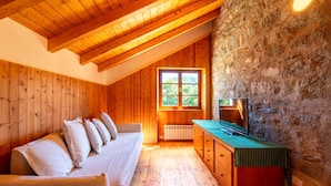Une pièce chaleureuse aux murs de bois, avec un plafond en pente, un mur en pierre, un canapé agrémenté de coussins, un téléviseur posé sur une table en bois et une fenêtre donnant sur un paysage verdoyant.