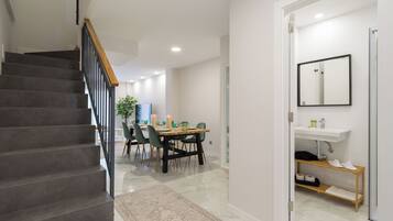 A modern interior with a staircase, a dining area with a wooden table and chairs, and a bathroom with a sink and mirror.
