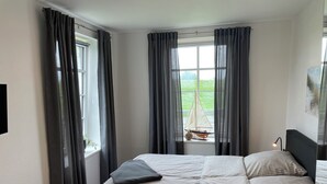 Et soveværelse med seng, et vindue med gardiner, en model af en sejlbåd og en lampe.