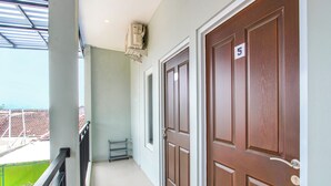 A hallway with two wooden doors, a metal handrail, a small metal shelf, and a window with a view of a green area outside. - OYO 92477 Anugrah Kostel Syariah (Semarang)