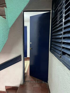 A narrow stairway leading to a dark blue door with a metal handle, flanked by a white wall and a window with horizontal blinds. - Departamentos Lomas SLP (San Luis Potosi)