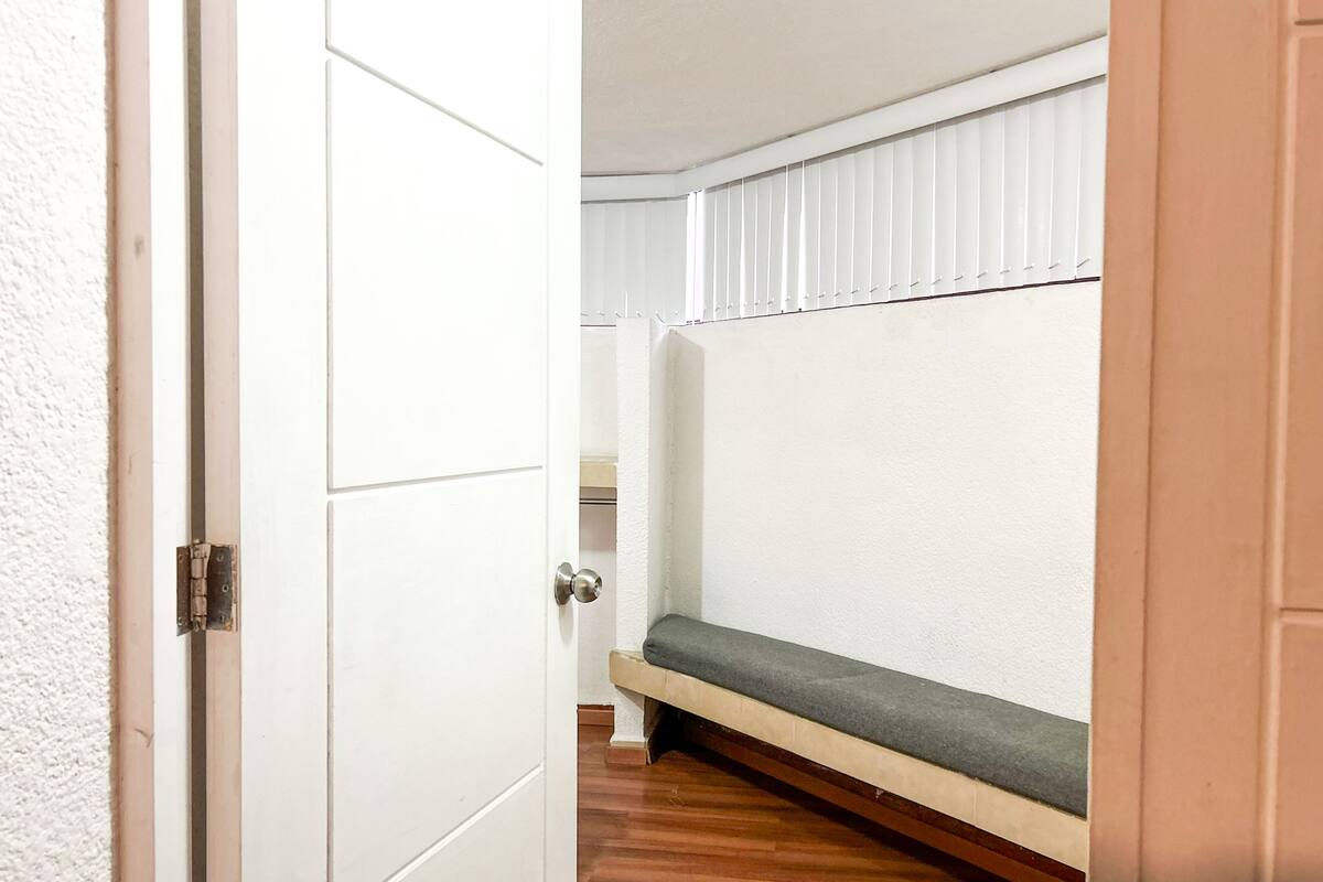 Un couloir étroit, doté d’un plancher en bois, mène à une pièce où se trouvent un banc et une fenêtre équipée de stores.