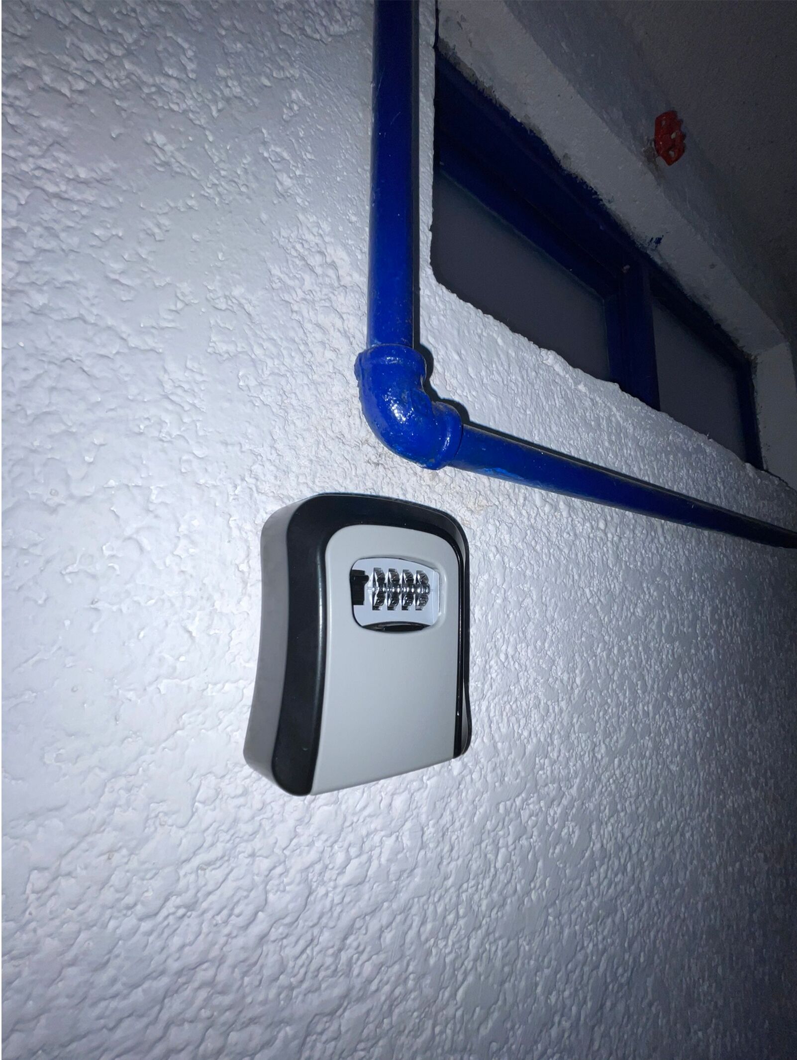 A white textured wall with a blue pipe and a small, rectangular device with a black and white label.