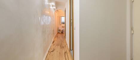 A hallway with wooden flooring and white walls, leading to a room with a window and a door.
