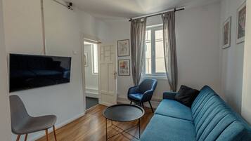 Una sala de estar con un sofá azul, un sillón gris, una mesita redonda y un televisor de pantalla plana montado en la pared.