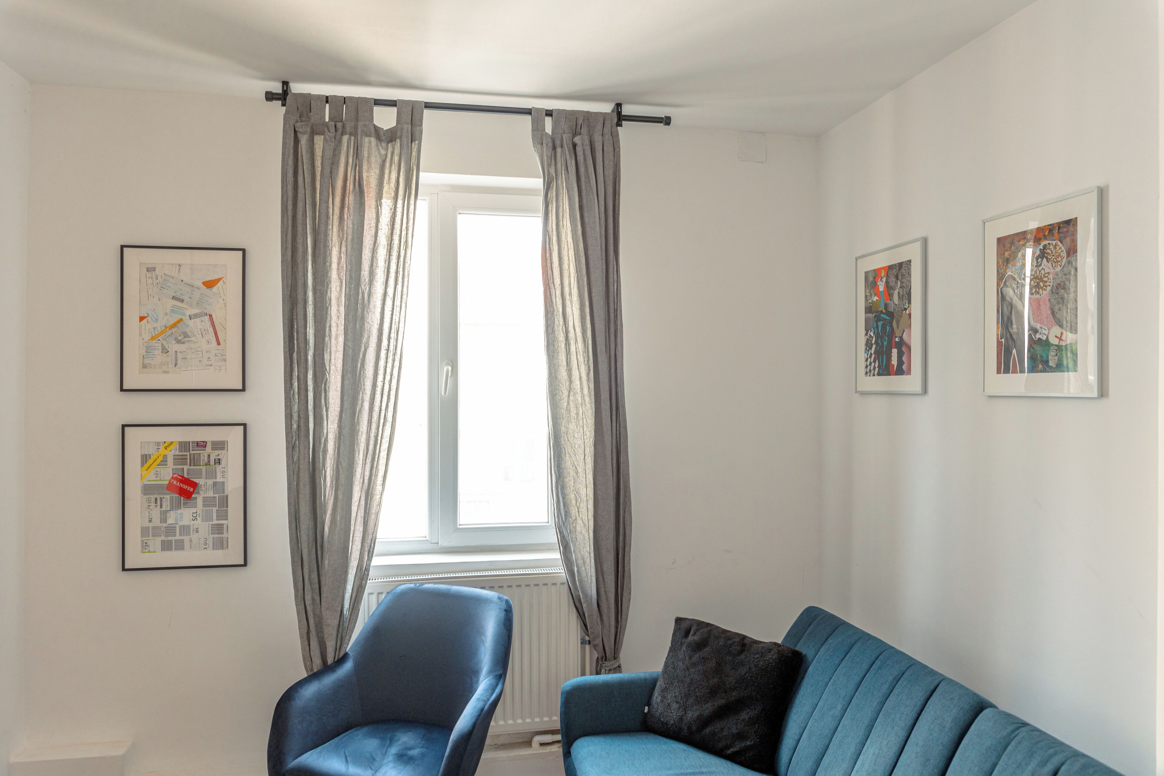 A living room with a blue sofa, a blue armchair, and a window with grey curtains. There are two framed pictures on the wall.