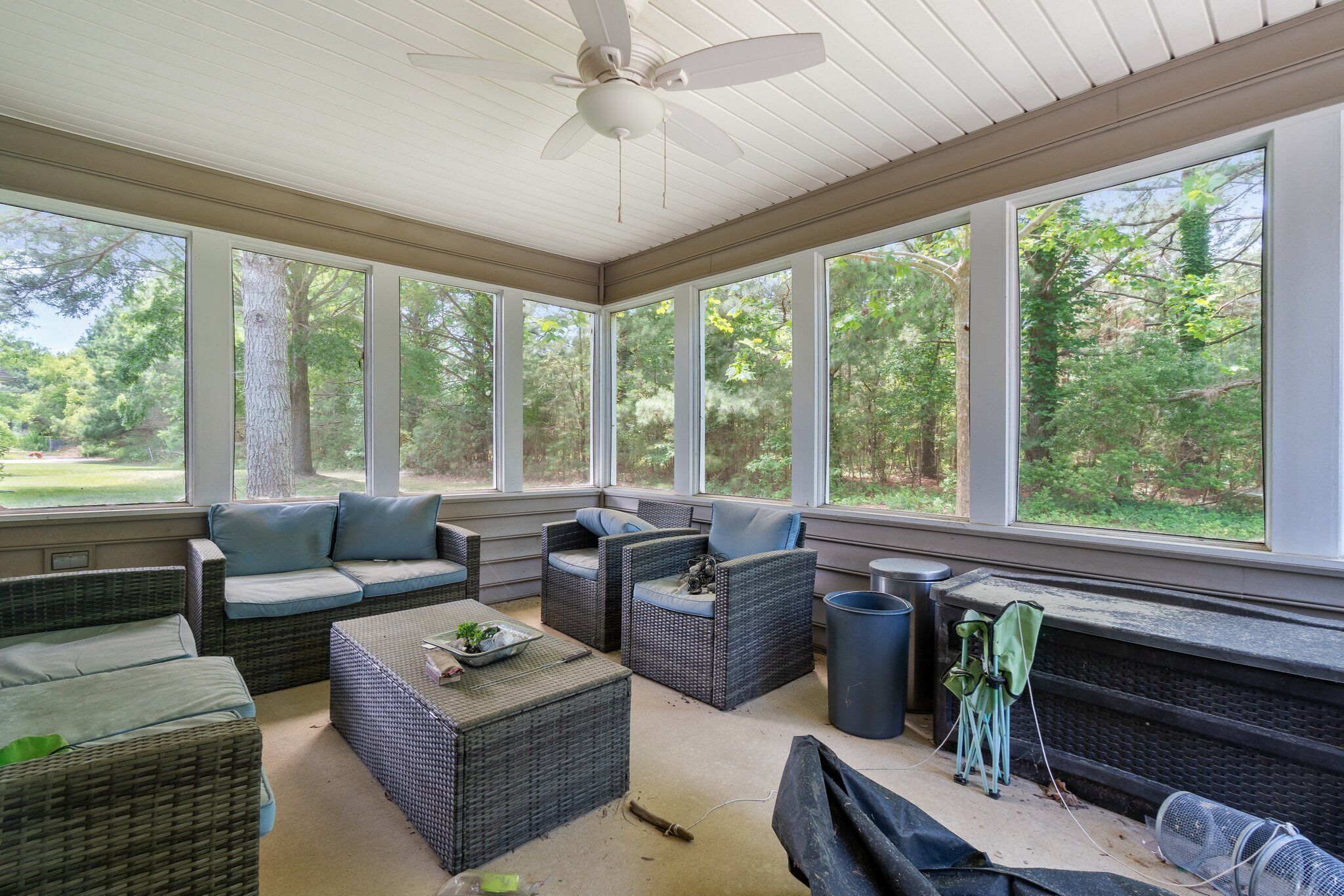 A covered outdoor seating area with wicker furniture, a ceiling fan, and large windows overlooking a wooded area.