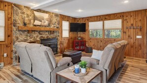 A cozy living room with a stone fireplace, wooden walls, a flat-screen TV, and a comfortable seating area. - “Bearly-Be-Haven” w/hot tub, firepit, Bearly-Be-Haven (Basye)