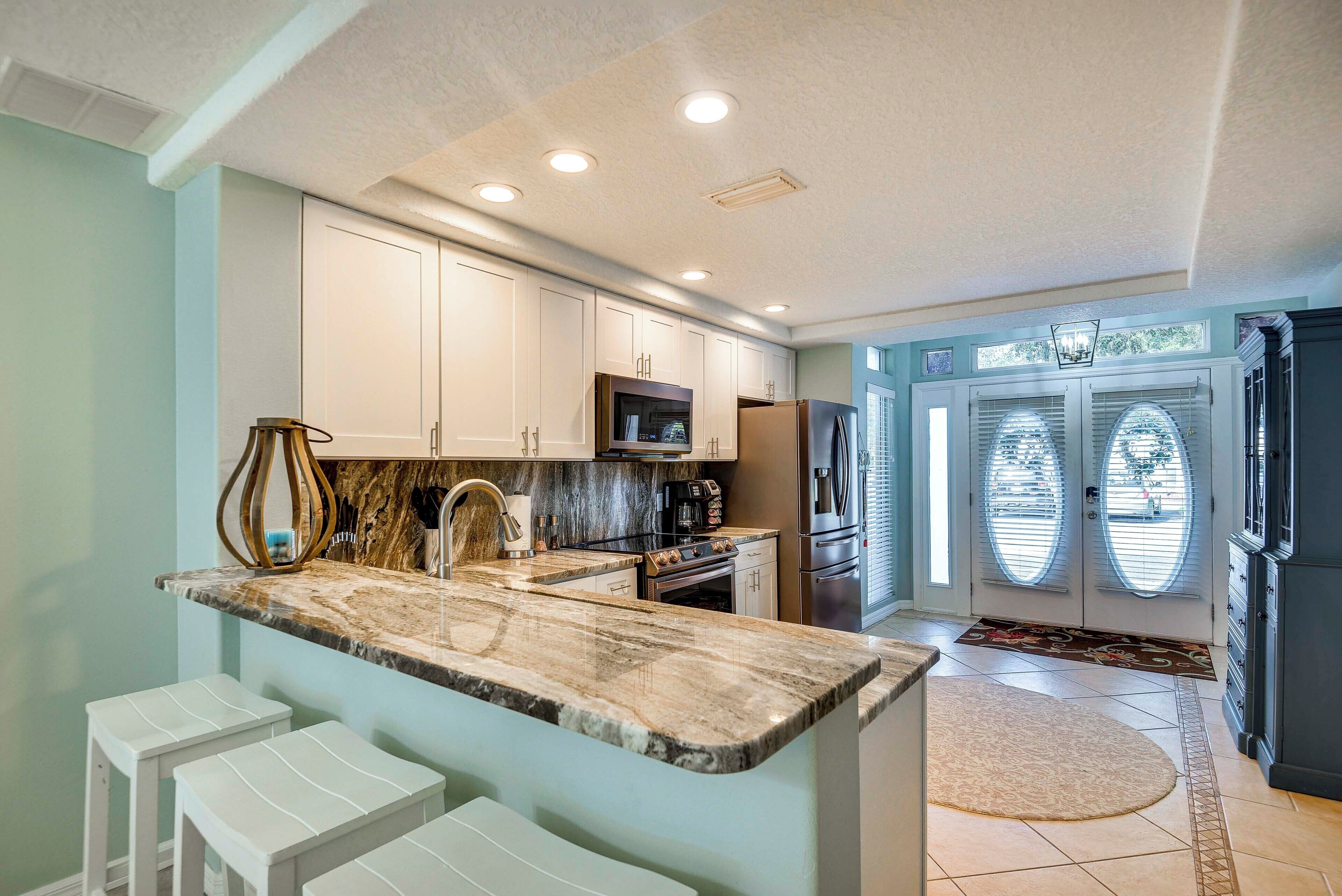 A kitchen with white cabinets, a marble countertop, and a built-in oven. There are three white bar stools and a large window with blinds.