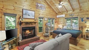 A cozy cabin interior with a fireplace, a bed, a television, and a ceiling fan.