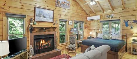 L’intérieur chaleureux d’un chalet avec une cheminée, un lit, un téléviseur et un ventilateur de plafond.