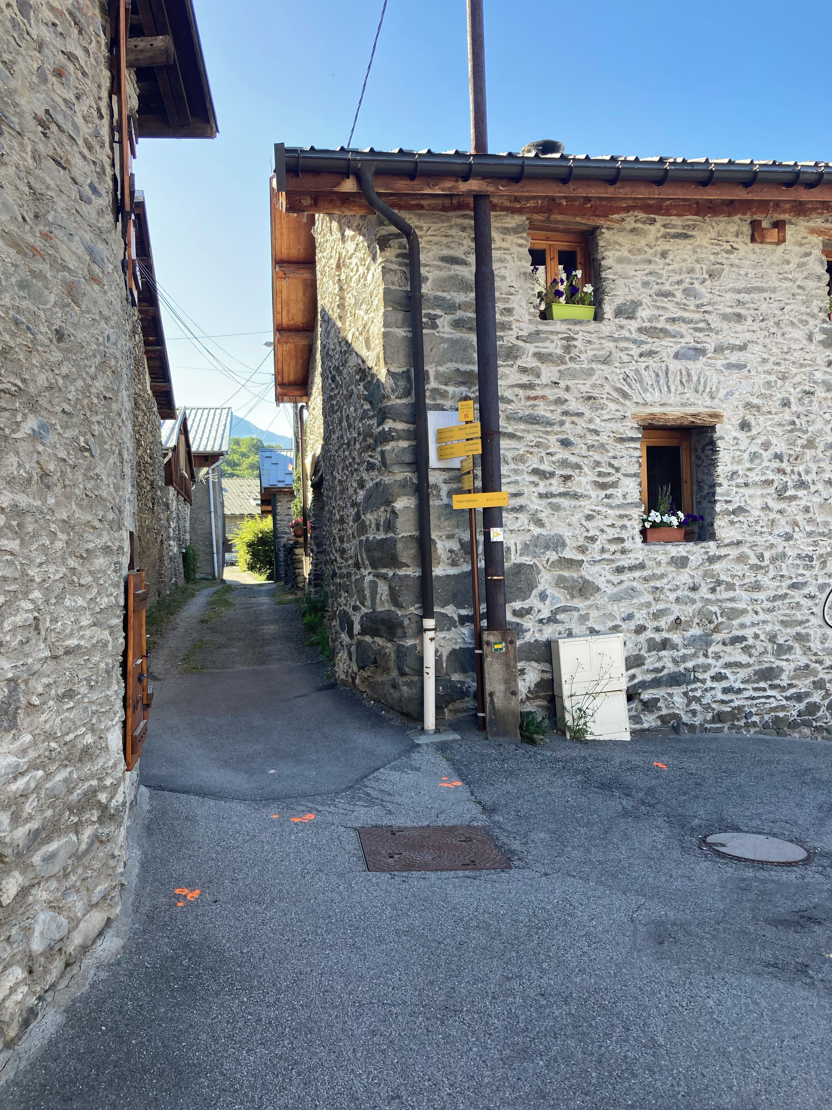 A narrow stone street with a wooden building on the right, featuring a small window with flowers and a signboard.