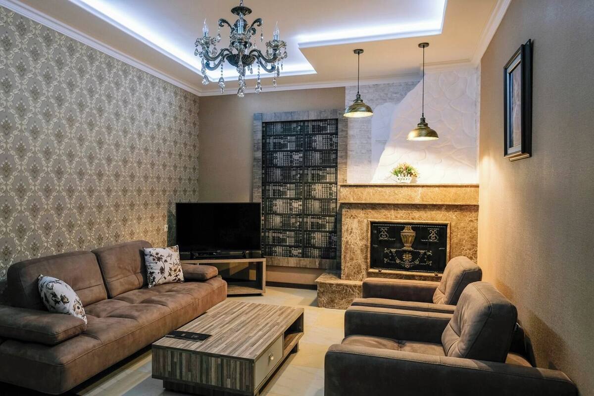 A living room with a sofa, armchair, coffee table, TV, bookshelf, fireplace, and chandelier.
