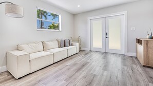 Een moderne woonkamer met een beige modulaire bank, een houten kast en een groot raam met uitzicht op palmbomen.